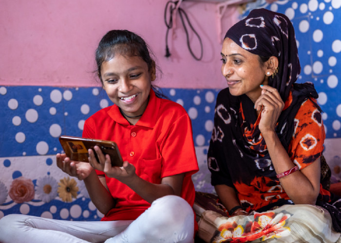 Mother with daughter sitting on bed sharing mobile phone screen
